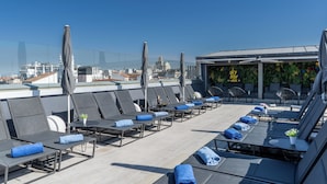 A rooftop lounge area with sun loungers, umbrellas, and a bar area with a view of the city skyline.