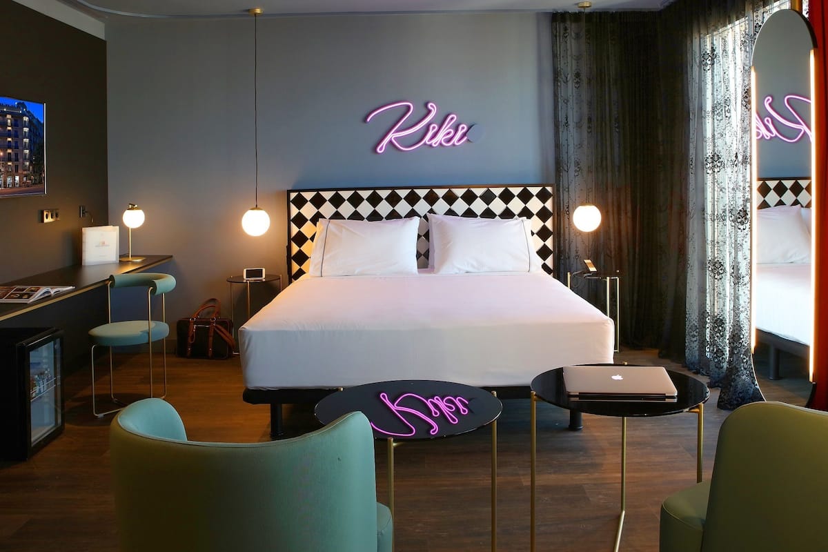 Habitación de hotel con cama, escritorio, silla, una computadora portátil y luces de neón en la pared.