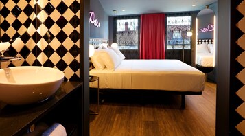 Habitación de hotel con una cama grande, una mesita de noche, un lavabo blanco y una ventana con cortina roja.
