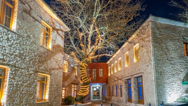 Un edificio de piedra con un árbol adornado con luces, un edificio rojo al fondo y un coche aparcado.