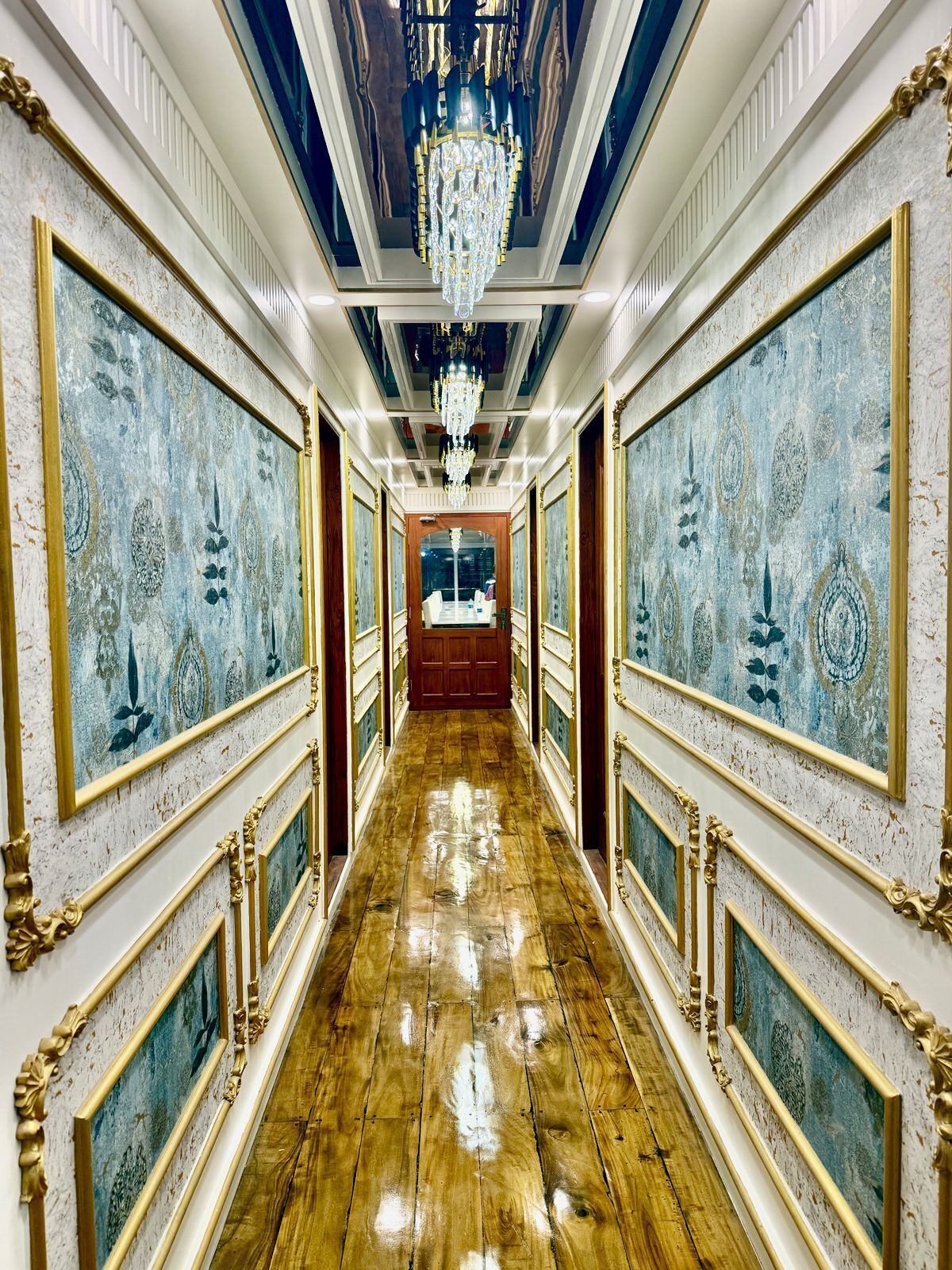 A hallway with ornate gold-framed paintings, a polished wooden floor, and a chandelier.