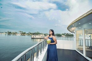 A person on a boat deck with a view of a river and distant buildings. - Sreekrishna Houseboats (Ambalapuzha)
