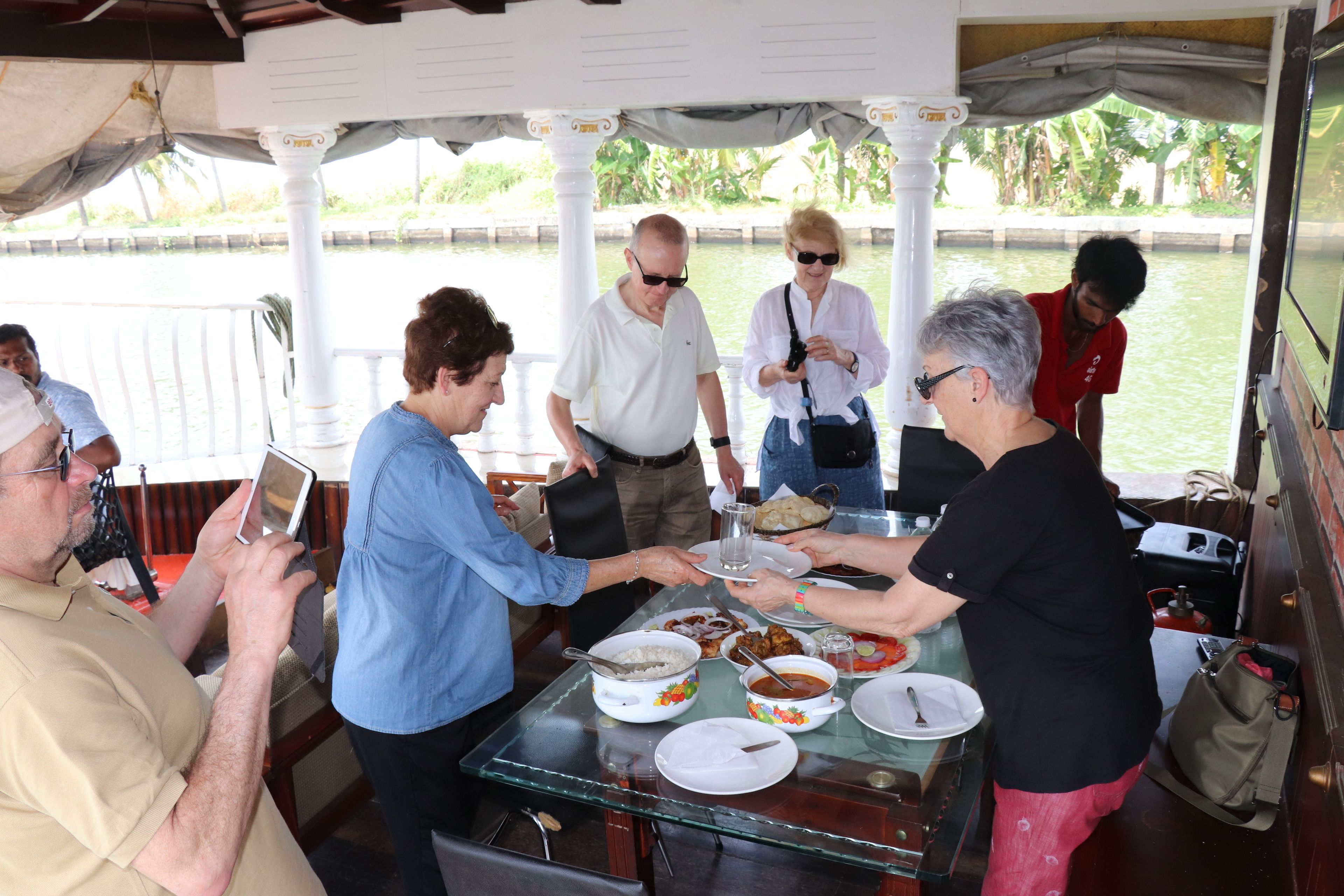 A group of people dining at a table with various dishes and a glass of water, under a covered area with columns.