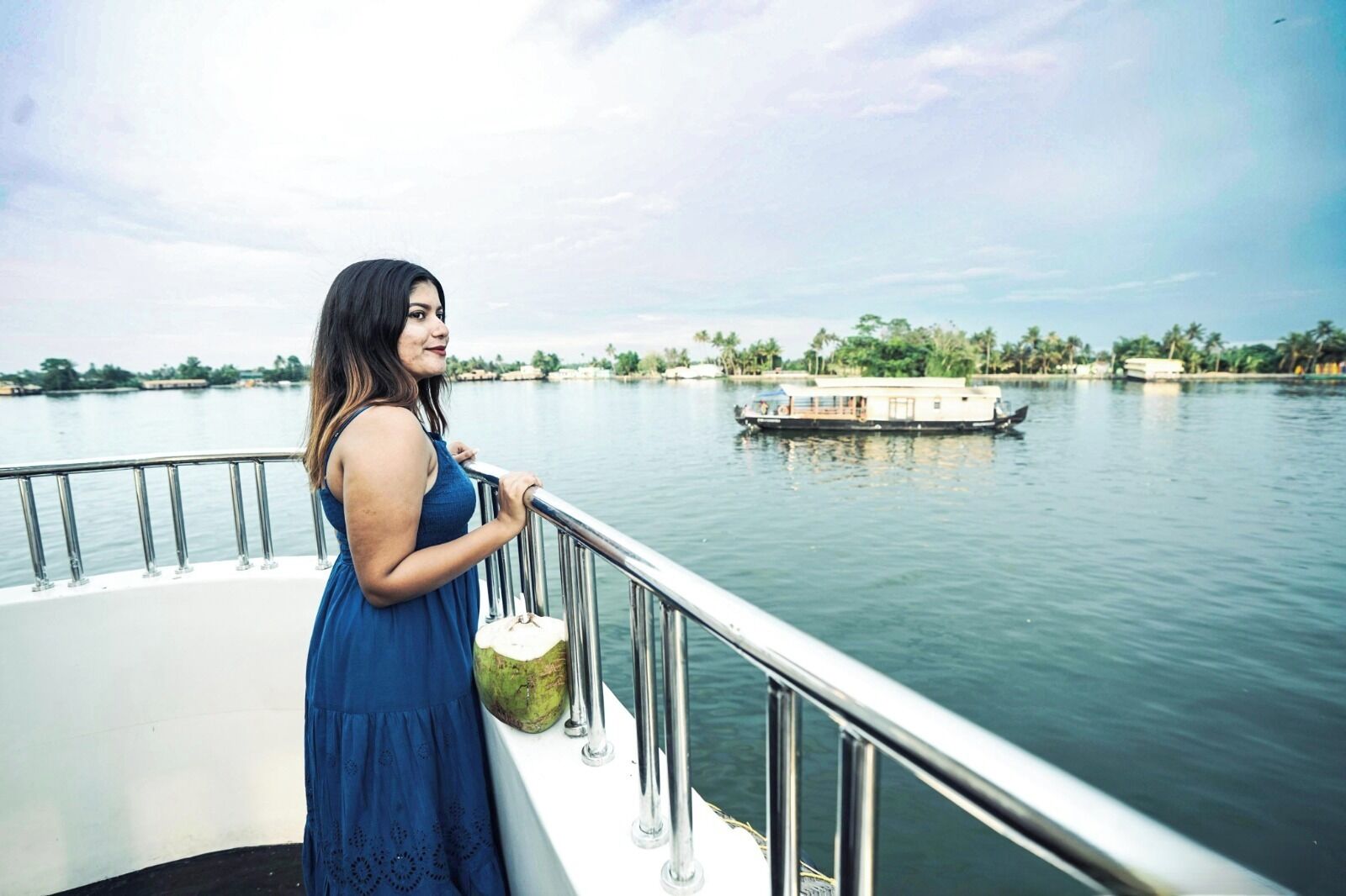 Una mujer con un vestido azul parada en un bote con barandilla metálica, mirando un río tranquilo donde se divisa una casa flotante a lo lejos.