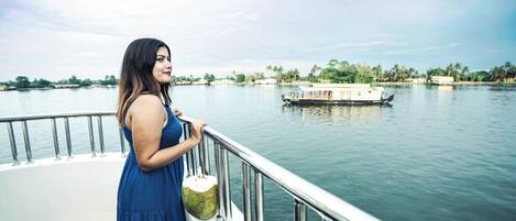 Una mujer con un vestido azul parada en un bote con barandilla metálica, mirando un río tranquilo donde se divisa una casa flotante a lo lejos.