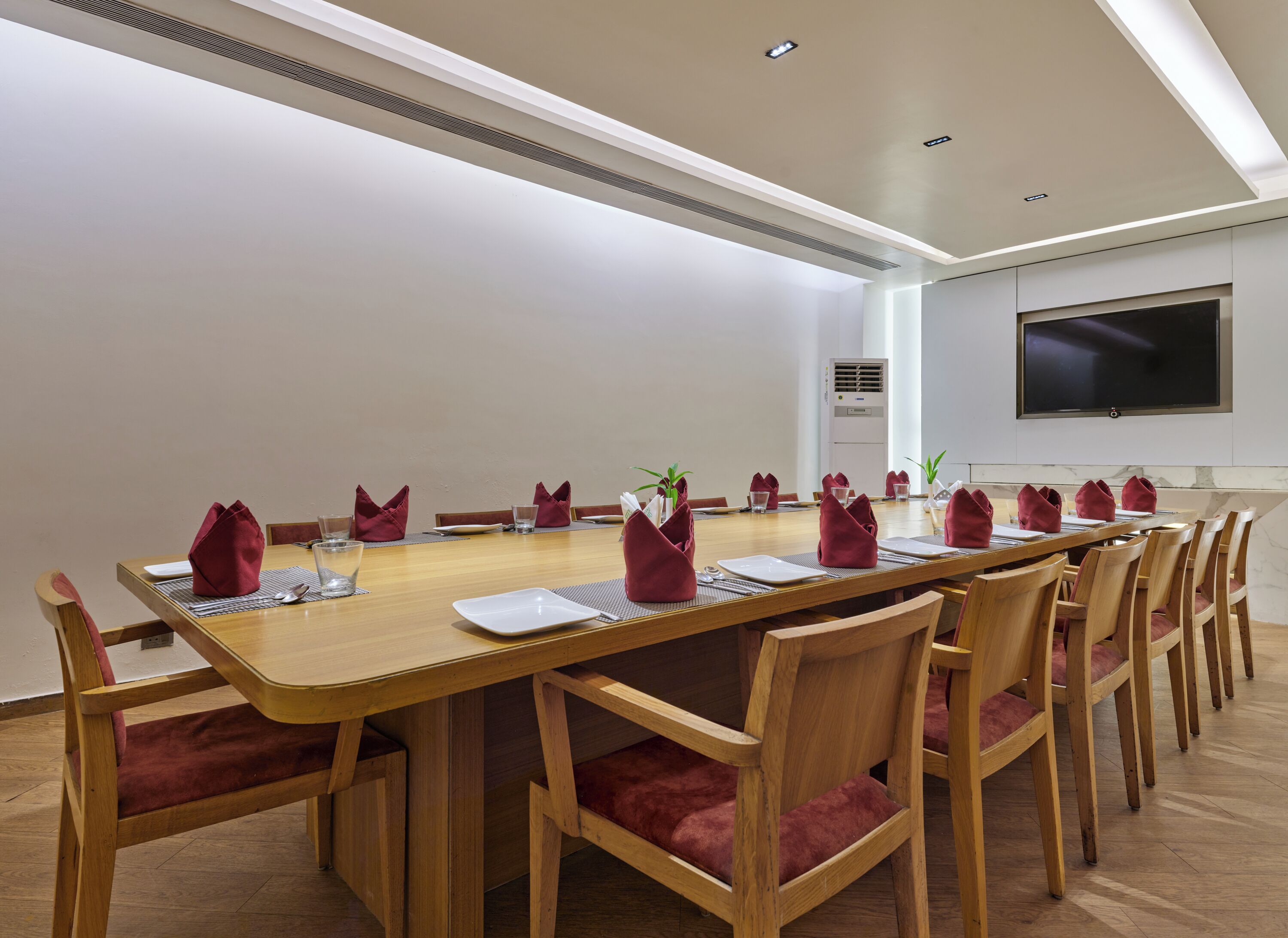 A conference room with a long wooden table, red upholstered chairs, and a flat-screen TV on the wall.