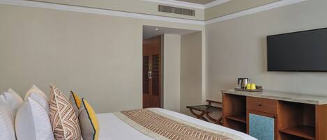 A hotel room with a large bed, a flat-screen TV mounted on the wall, a wooden desk with a chair, and a small kitchenette area.