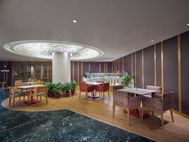 En modern hotellobby med en central, cirkelformad lampa, en sittgrupp med trä- och mönstrade stolar, ett marmorgolv och ett bord med efterrätter i bakgrunden.