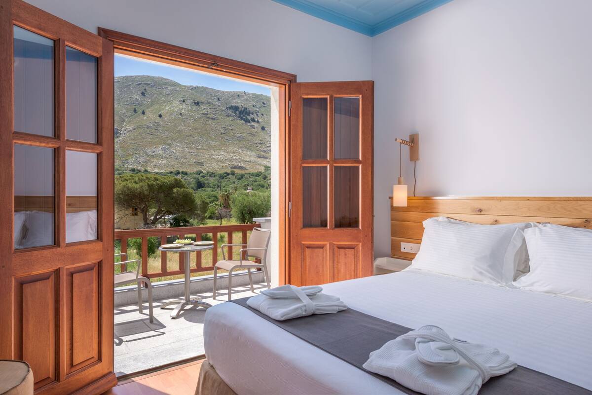 Un dormitorio con una cama grande, una puerta de madera que da a un balcón y vistas a un paisaje montañoso.