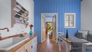 Una cocina con encimera de madera, un fregadero y una estantería con vajilla y vasos. Hay una zona de comedor con una mesa y sillas, y una ventana con vistas al exterior.
