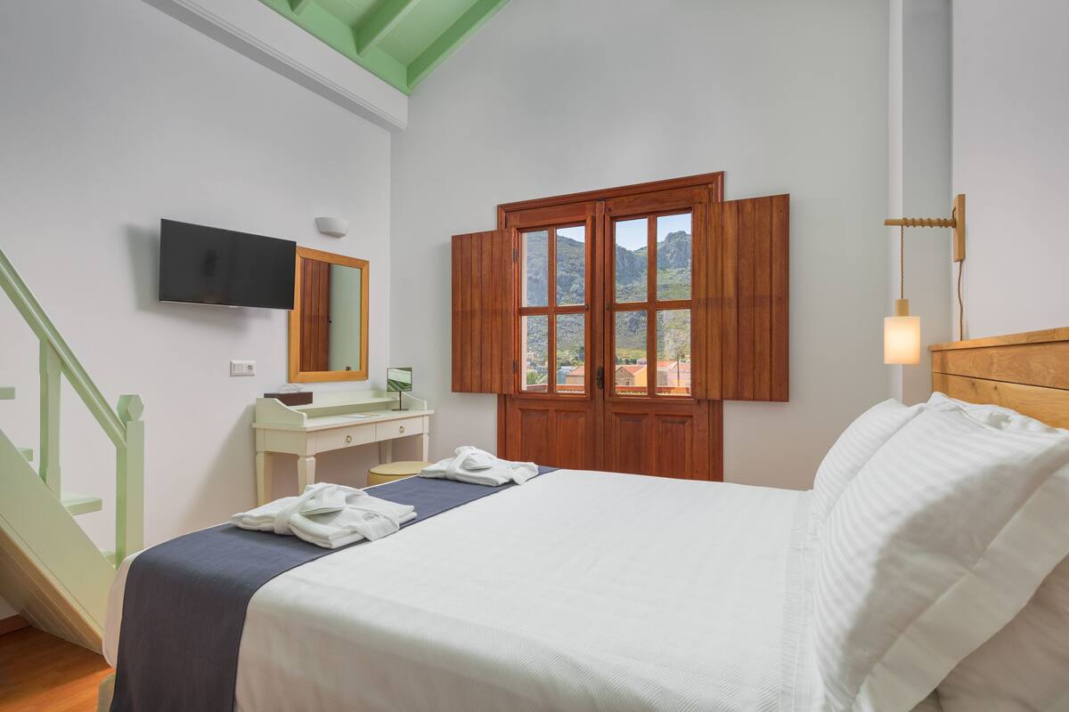 Un dormitorio con cama, escritorio, espejo, televisión y ventana con persianas de madera.