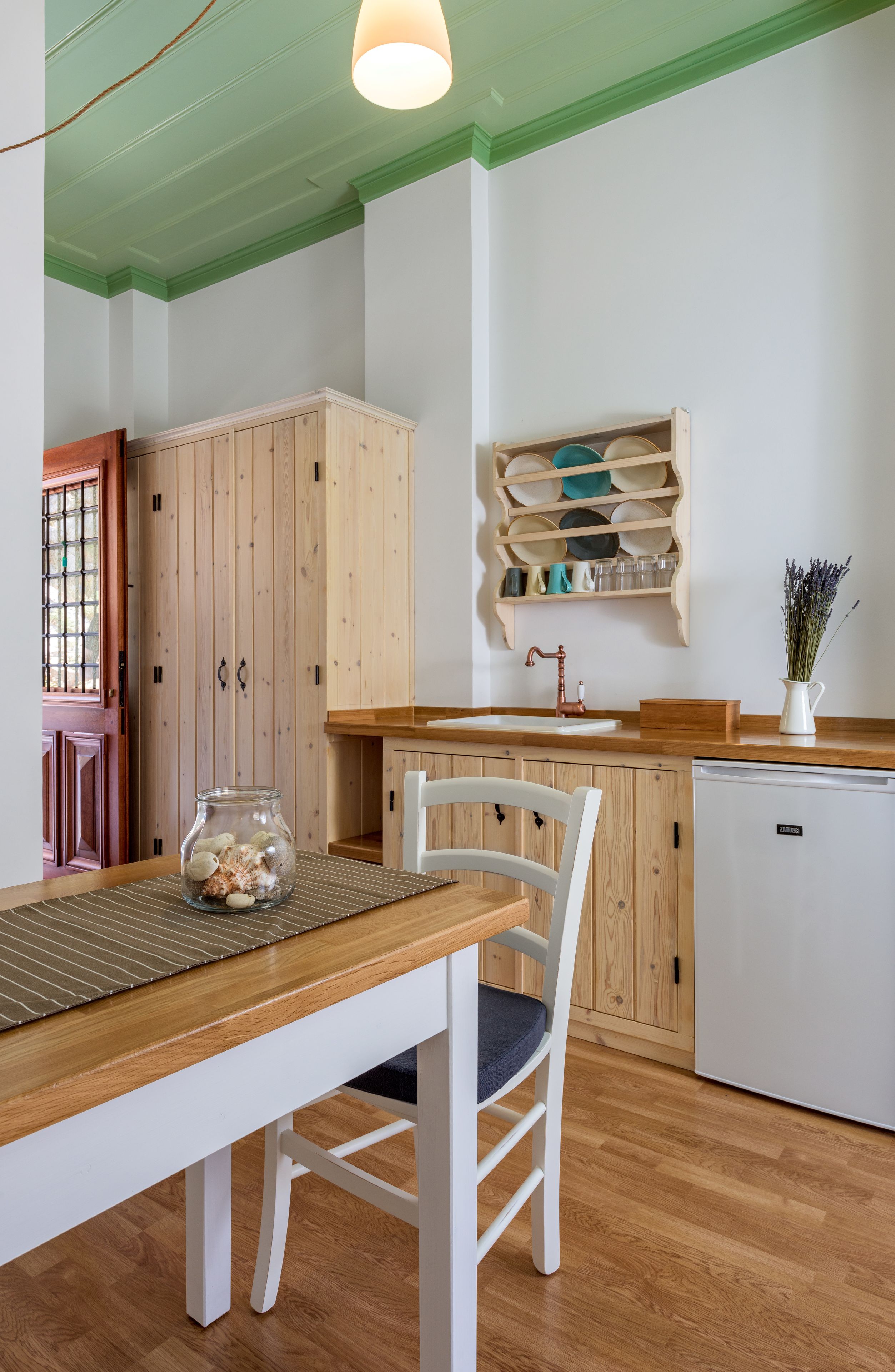 Una cocina con armarios de madera, un refrigerador blanco, una mesa de comedor de madera y una silla, además de un escurreplatos colgante.