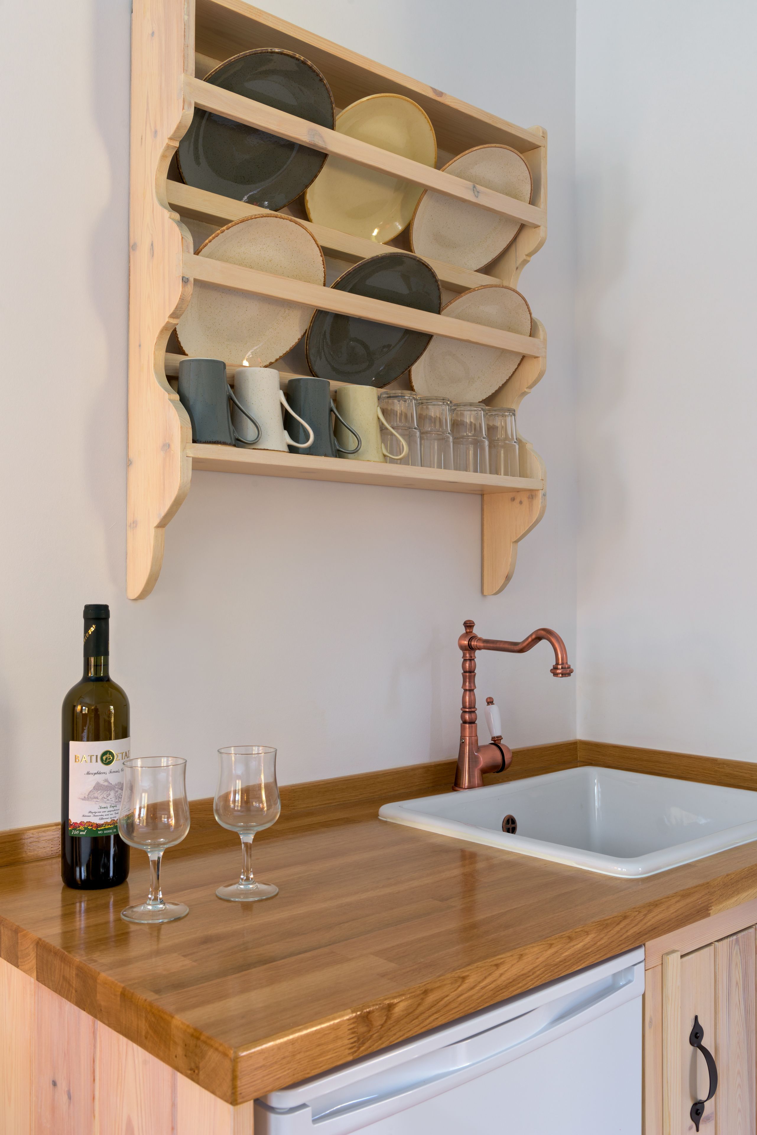 Una cucina con piano di lavoro in legno, lavello bianco, rubinetto in rame, una bottiglia di vino e un espositore per piatti e bicchieri.