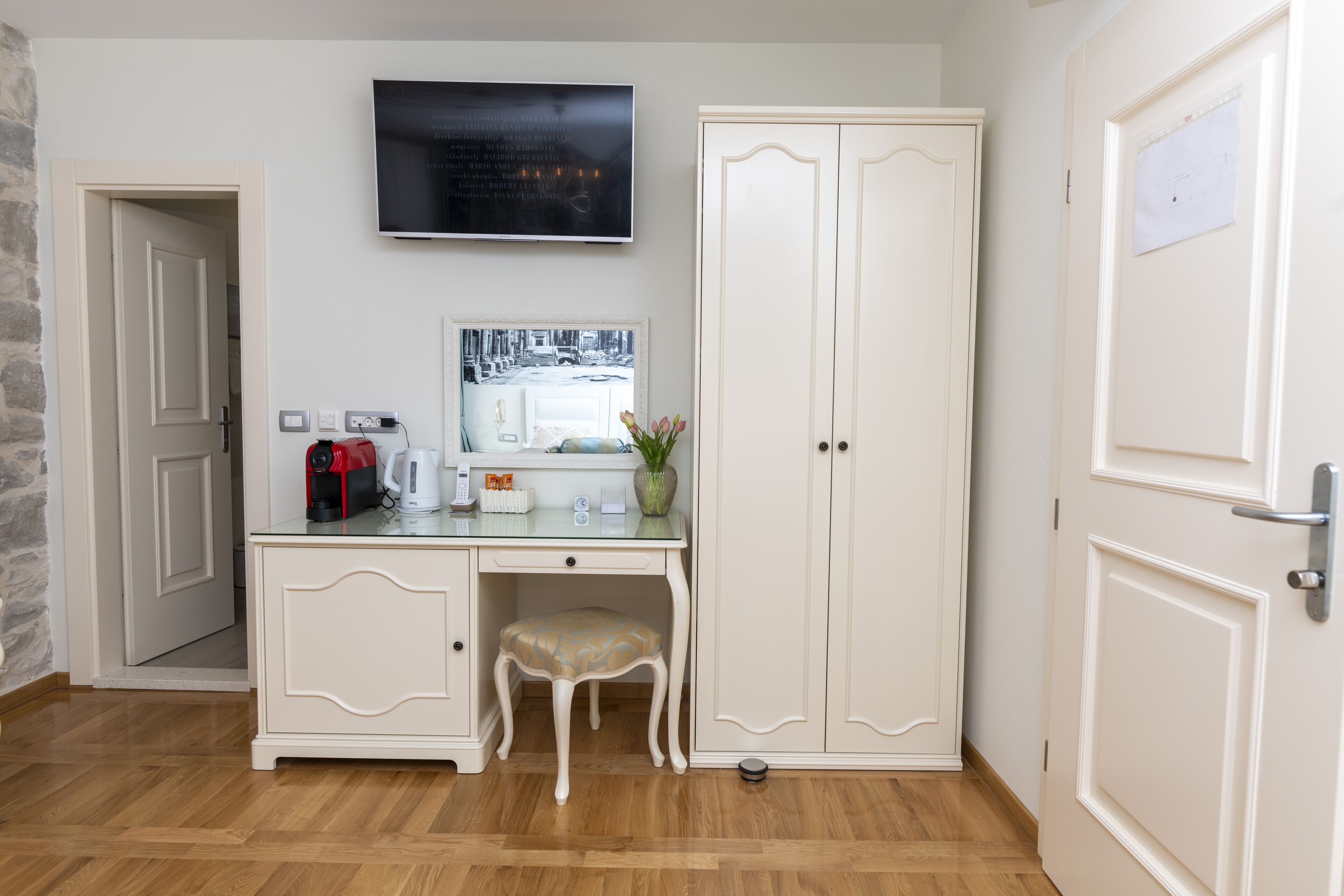 Una habitación con piso de madera, un escritorio blanco con una silla, un televisor y un armario.