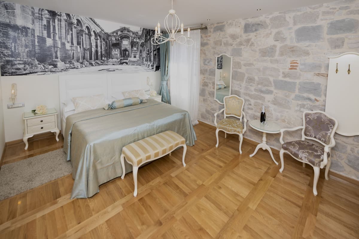 A bedroom with a large bed, a bench, a chair, a small table, and a mirror on a stone wall.