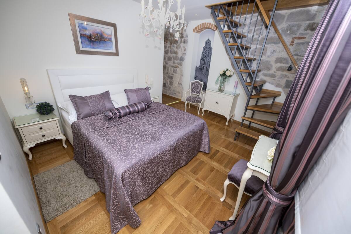 A bedroom with a bed, nightstand, chair, and a staircase.