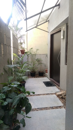 A narrow outdoor corridor with potted plants, a concrete pathway, and a door with a metal handle.