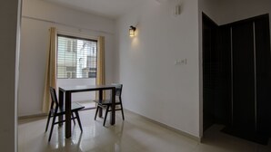 A room with a dining table and chairs, a window with curtains, a wall-mounted light, and a black wardrobe.