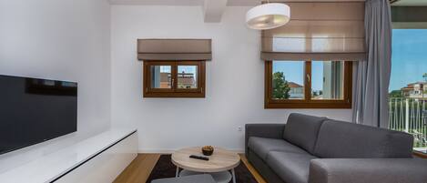 Una sala de estar moderna con un televisor de pantalla plana, un sofá gris, una mesita redonda con un bol y una ventana con vistas a edificios.