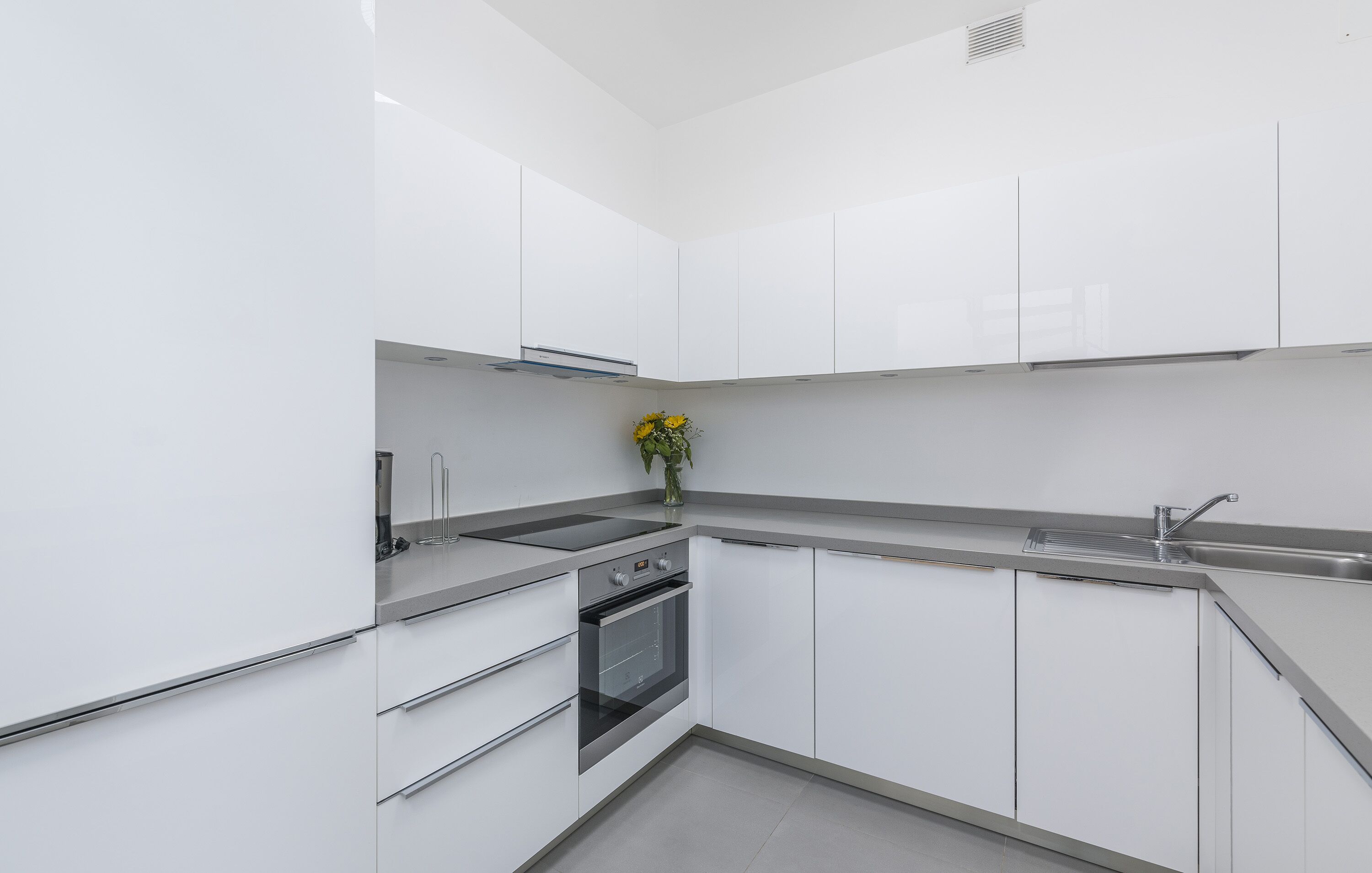 Een moderne keuken met witte kasten, een inbouwoven, een roestvrijstalen spoelbak en een vaas met gele bloemen op het aanrecht.