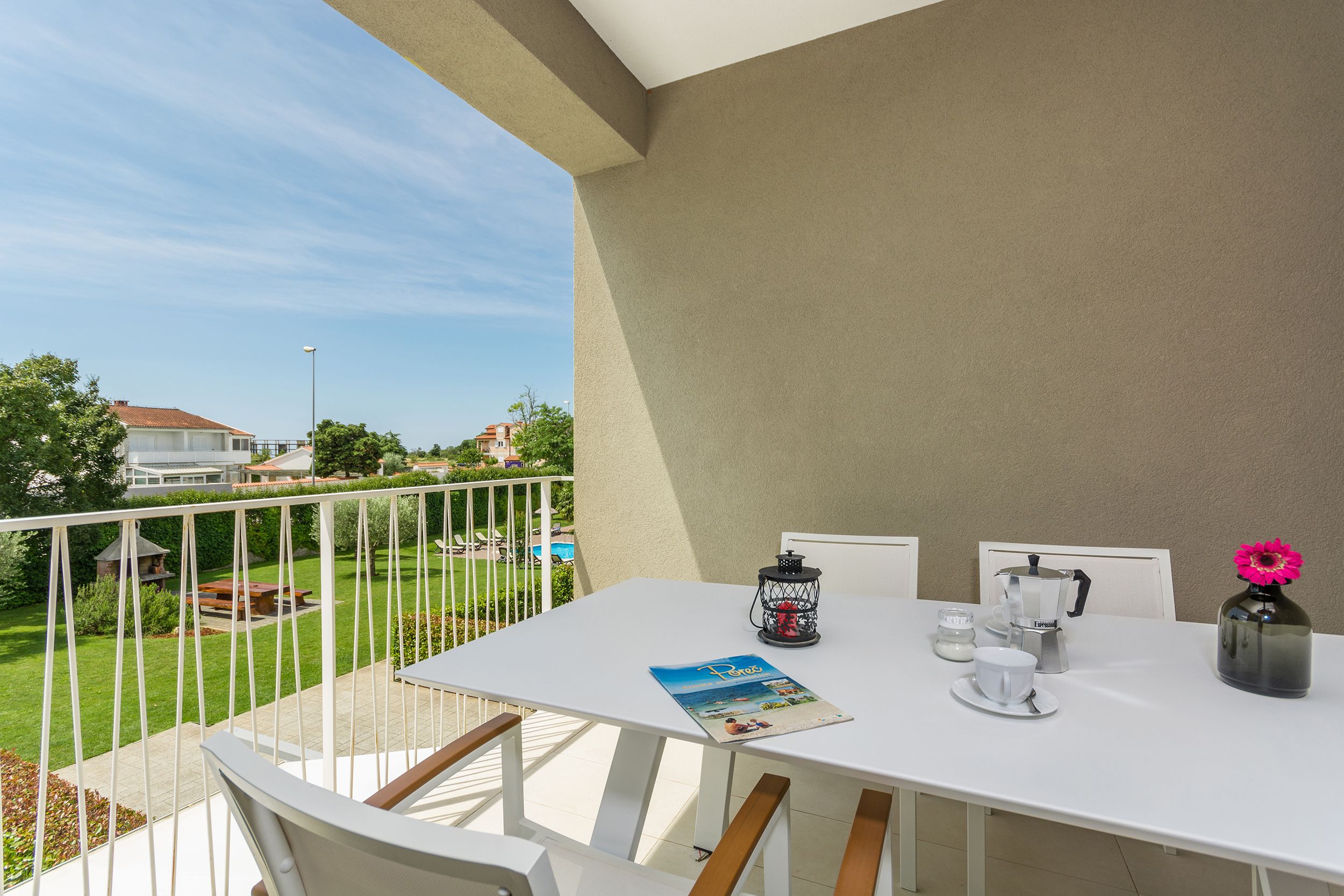 Un balcon avec une table blanche, des chaises, un magazine, une machine à café, une tasse et un vase avec une fleur.