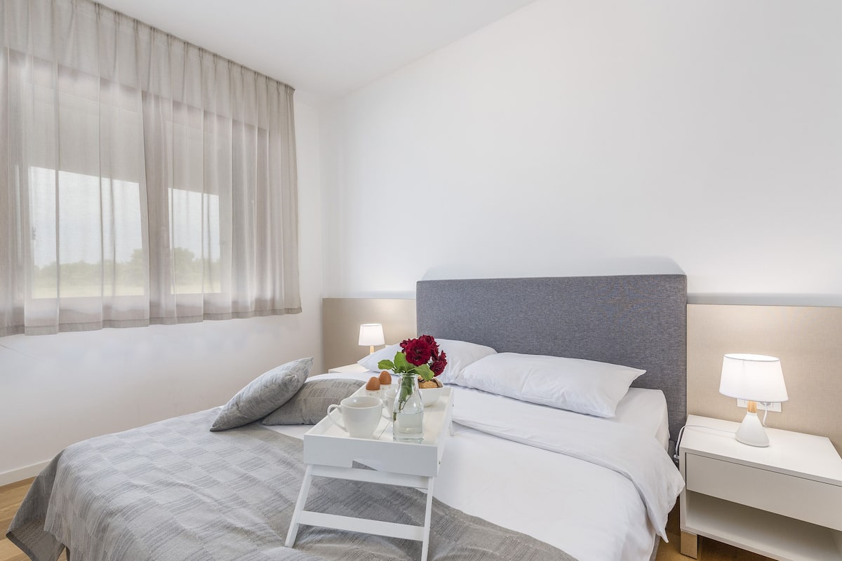 A modern hotel room with a large bed, a bedside table with a lamp, and a window with sheer curtains.
