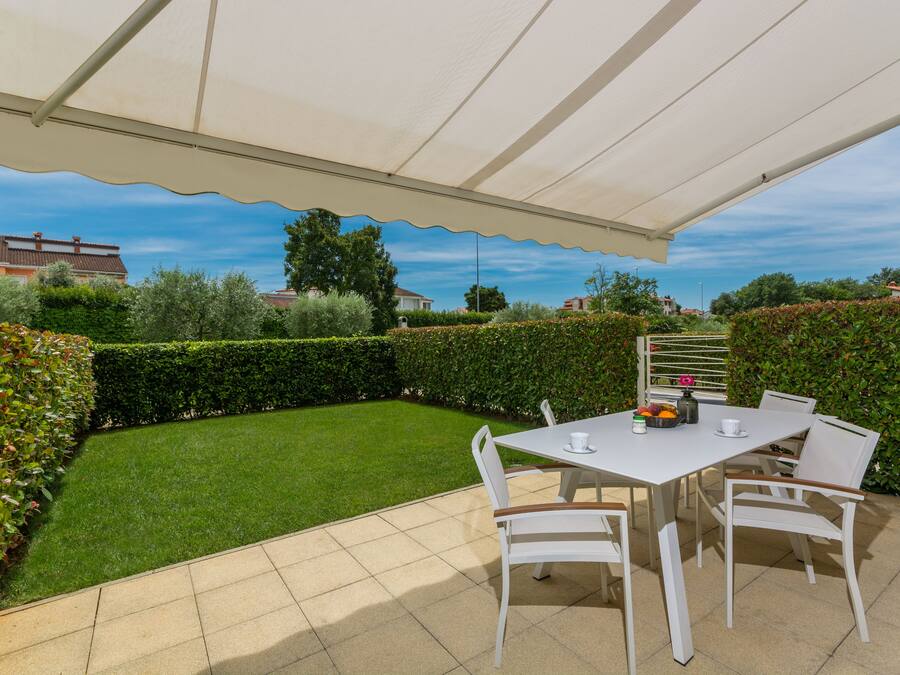 Un patio exterior con una mesa y sillas, rodeado de un césped cuidado y setos.