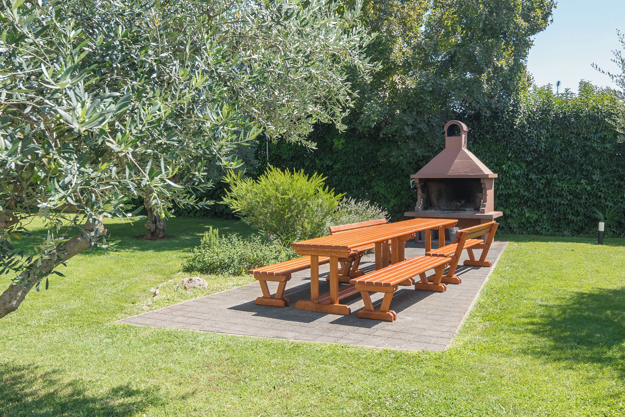 A picnic area with a wooden table and benches, surrounded by greenery and a brick patio.