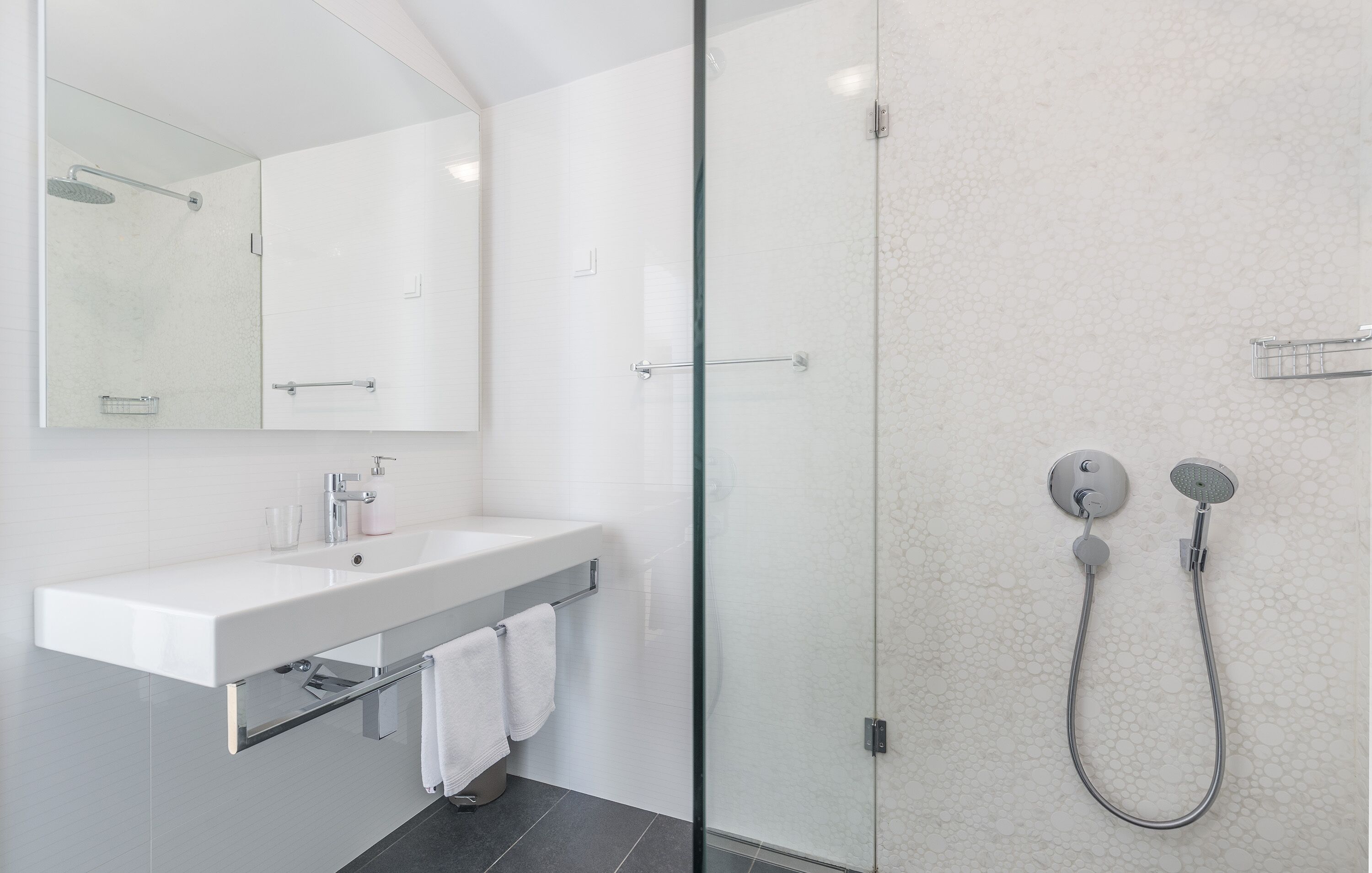 Un baño moderno con ducha envuelta en vidrio, un lavamanos blanco con espejo superior y un perchero para toallas.