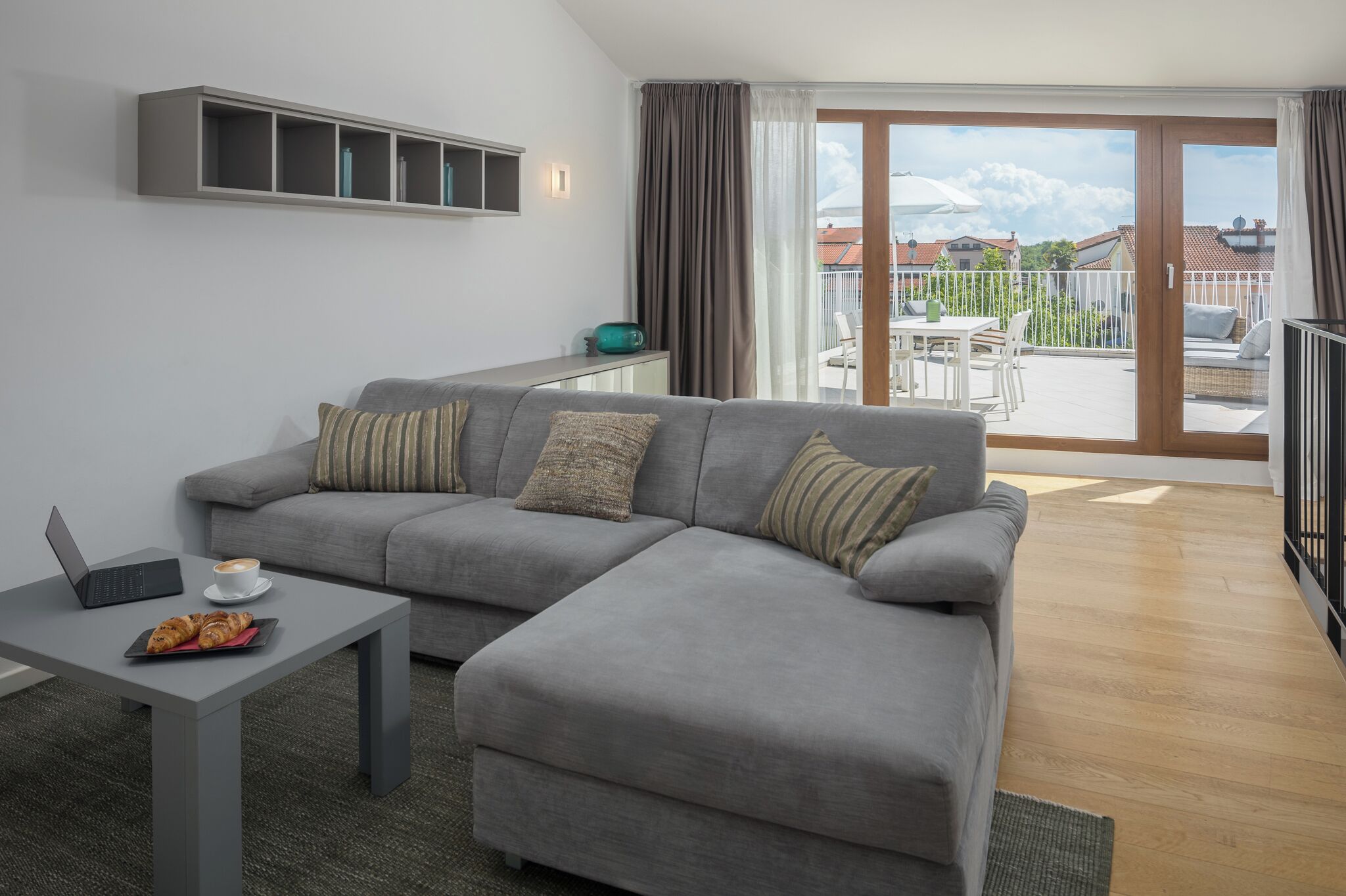 A modern living room with a grey sofa, a coffee table, and a laptop. There is a balcony with a view of houses and a clear sky.