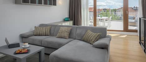 A modern living room with a grey sofa, a coffee table, and a laptop. There is a balcony with a view of houses and a clear sky.
