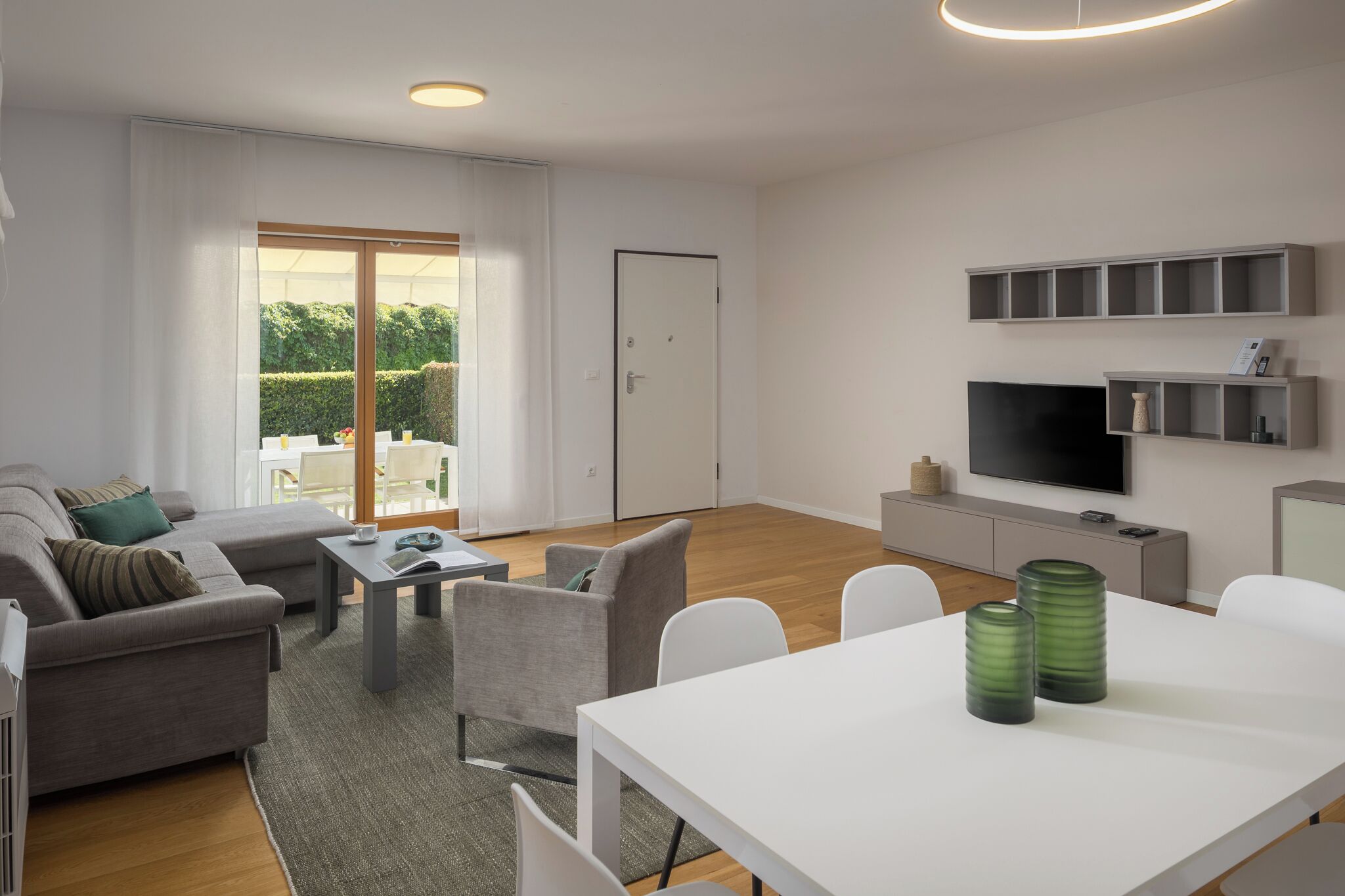 A modern living room with a sofa, chairs, a dining table, a TV, and a sliding glass door to a garden.
