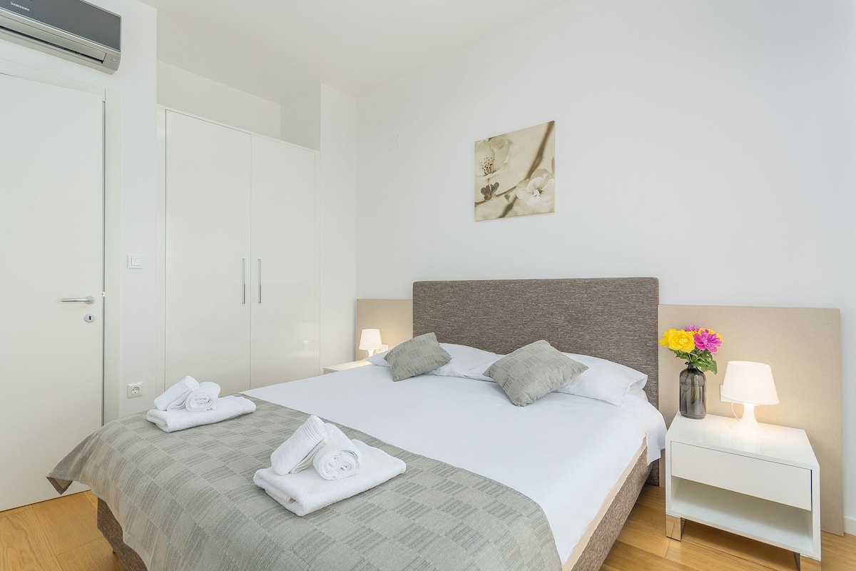 A hotel room with a neatly made bed, a bedside table with a lamp, a wall-mounted air conditioner, and a framed picture on the wall.