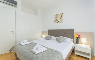 Una habitación de hotel con una cama bien hecha, una mesita de noche con lámpara, aire acondicionado en la pared y un cuadro enmarcado.
