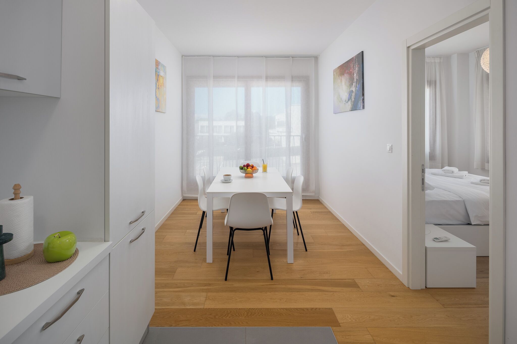 Een moderne, minimalistische kamer met een witte eettafel, stoelen en een klein keukentje met kastjes en een groene appel op het aanrecht.