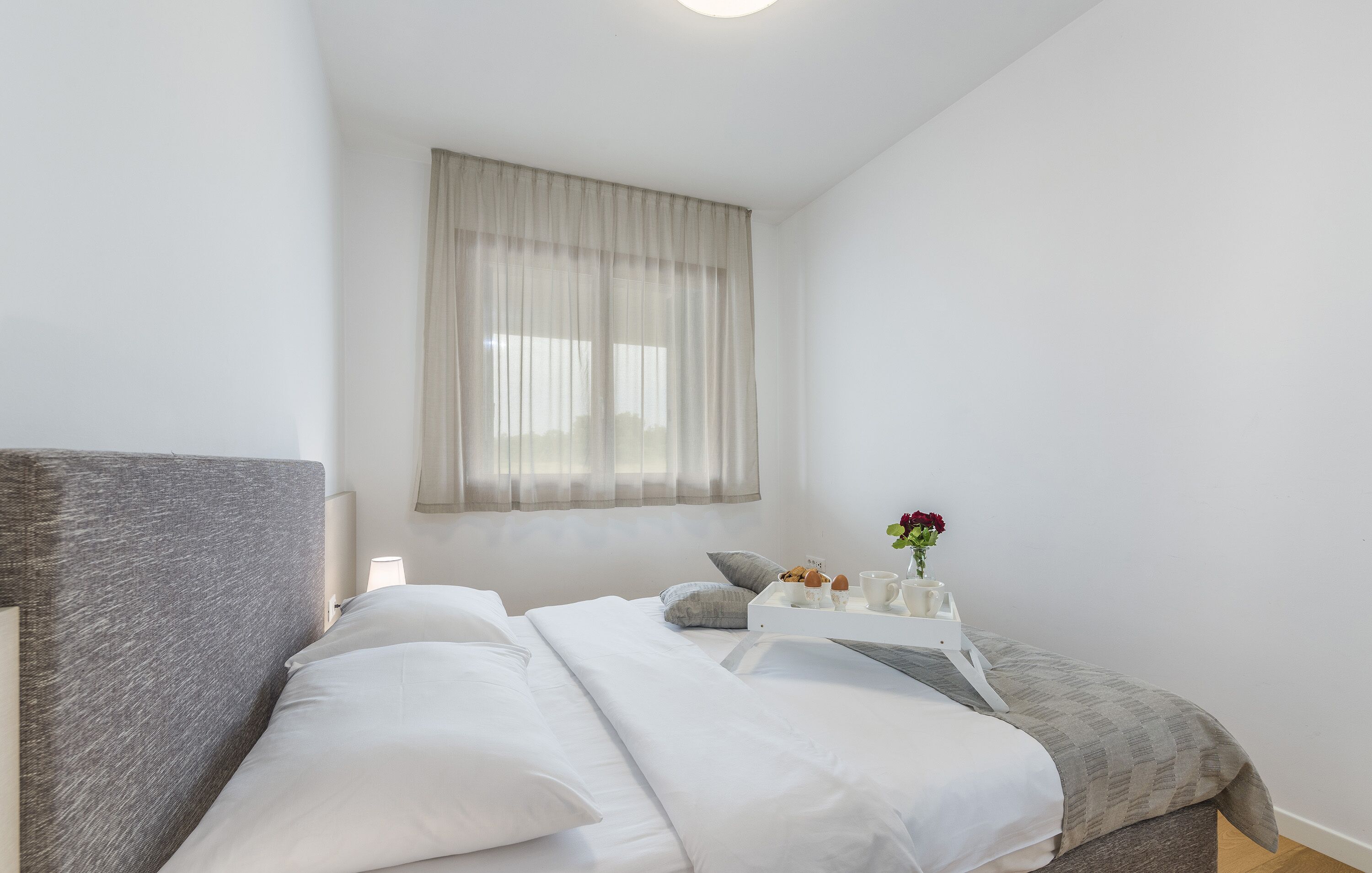 A bedroom with a bed, pillows, a bedside table with a vase of flowers, and a window with sheer curtains.