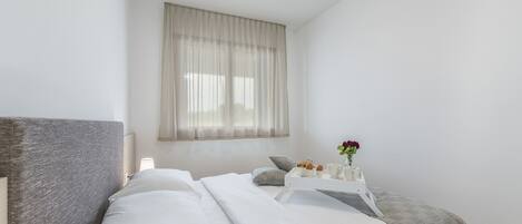 A bedroom with a bed, pillows, a bedside table with a vase of flowers, and a window with sheer curtains.