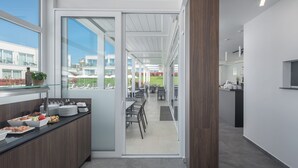 A modern hotel lobby with a glass partition, a buffet area with food and drinks, and a view of the outdoor garden. - Apartments Maj Residence Premium (Porec)