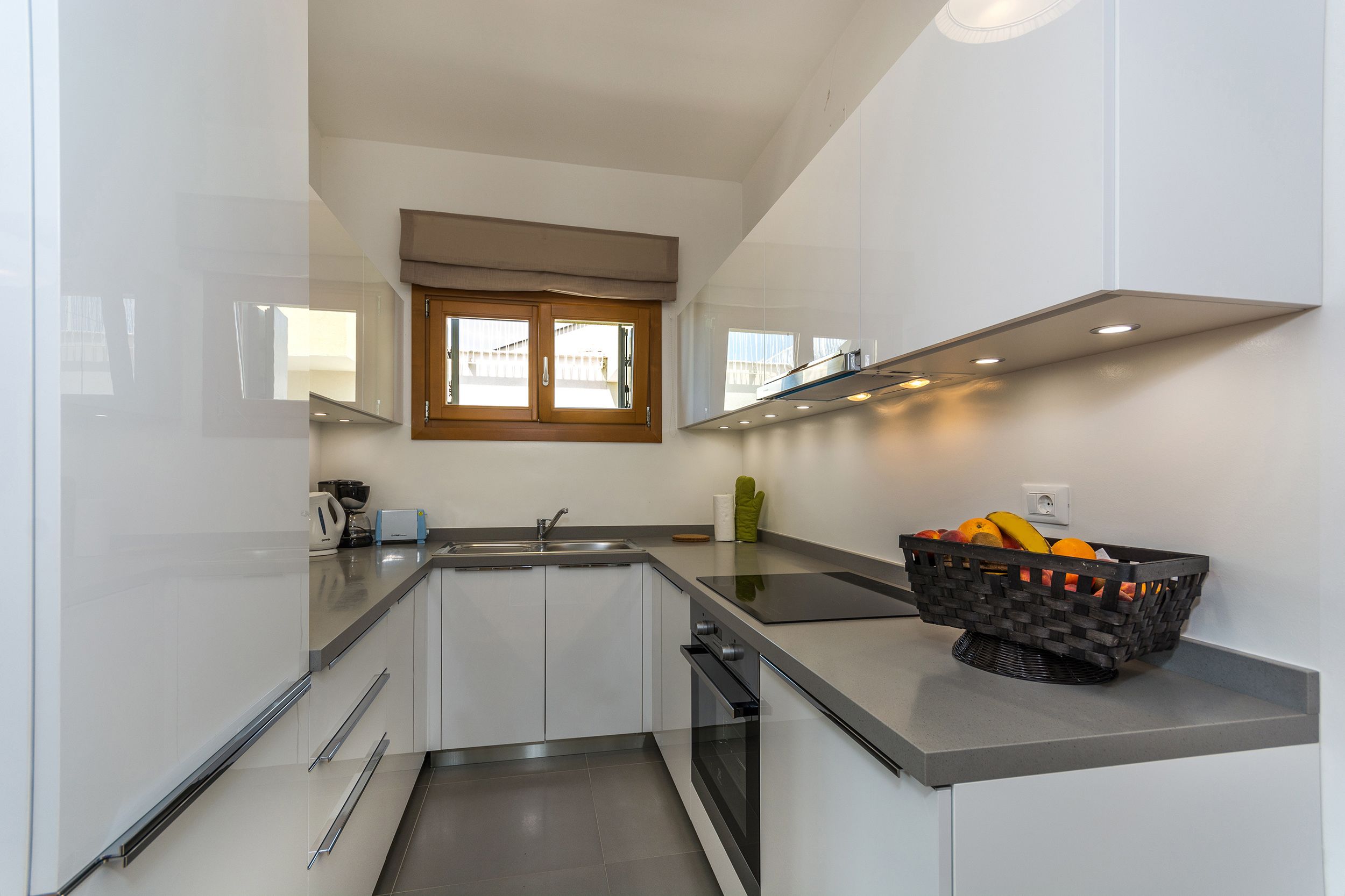 Una cocina moderna con armarios blancos, encimera negra, ventana con persianas y una cesta de frutas.
