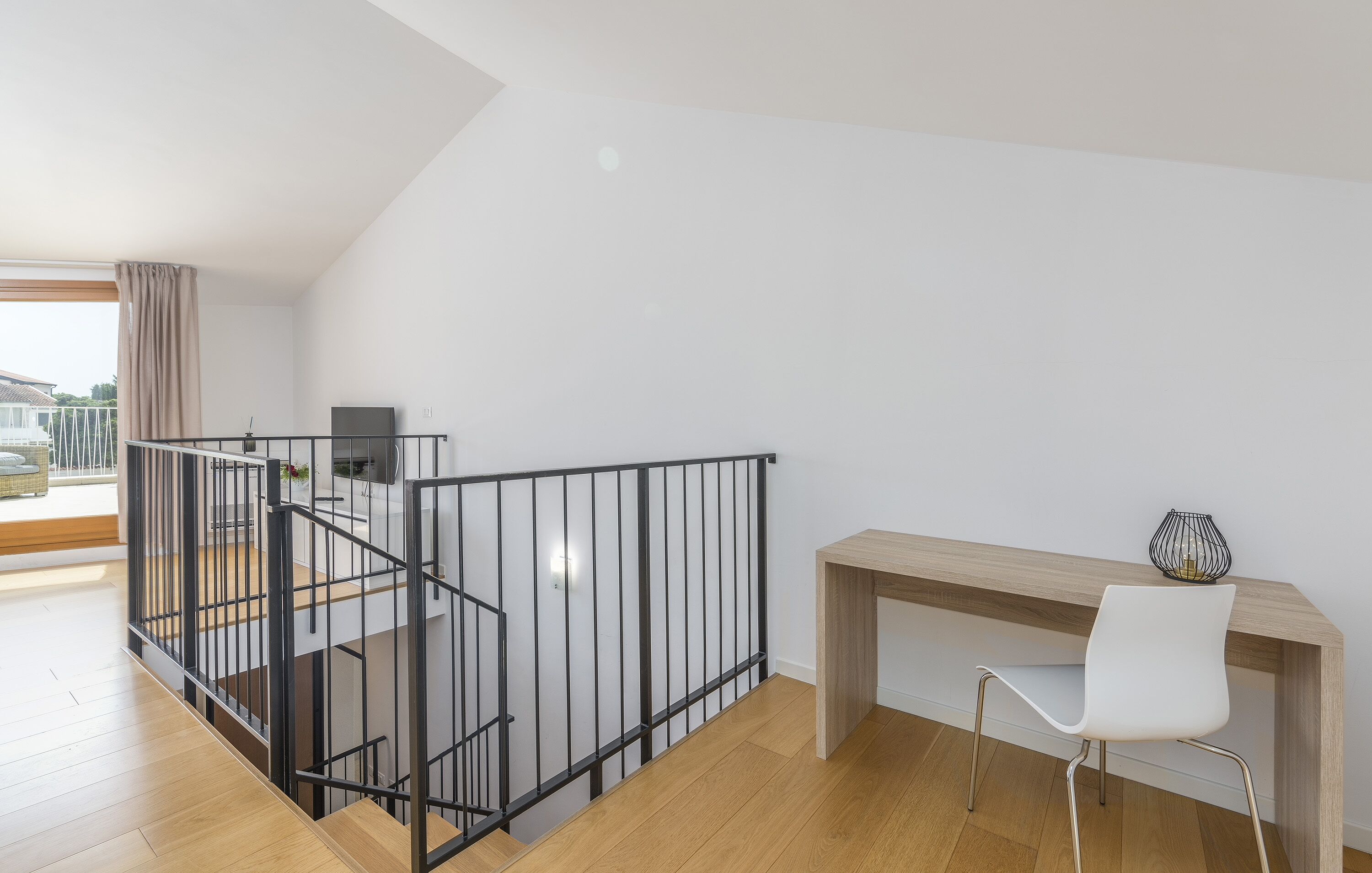 A modern apartment with a wooden floor, a black metal railing, a white chair, and a wooden desk.
