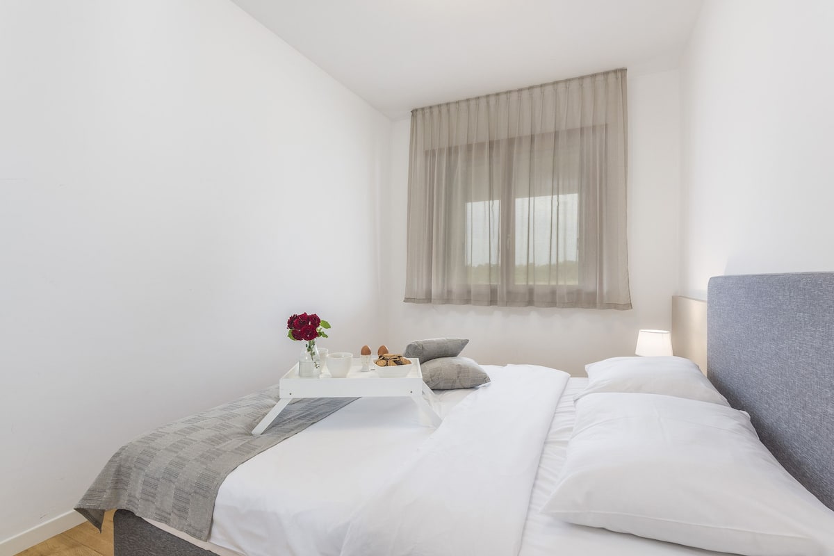 A neatly arranged bedroom with a bed, bedside table, and a window with sheer curtains.