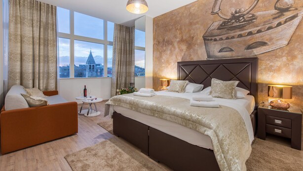 A modern hotel room with a large bed, a sofa, a small table, and a view of a cityscape through large windows. - PRIMA Life Spalato (Split)
