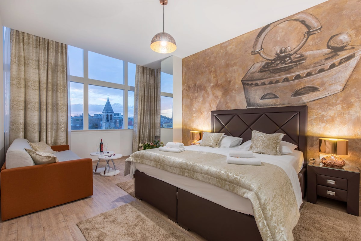 Una habitación de hotel moderna con una cama grande, un sofá, una mesita y vistas a la ciudad a través de amplias ventanas.