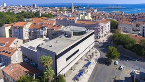 An aerial view of a city with a prominent modern building with a flat roof and a large open plaza in front. - PRIMA Life Spalato (Split)