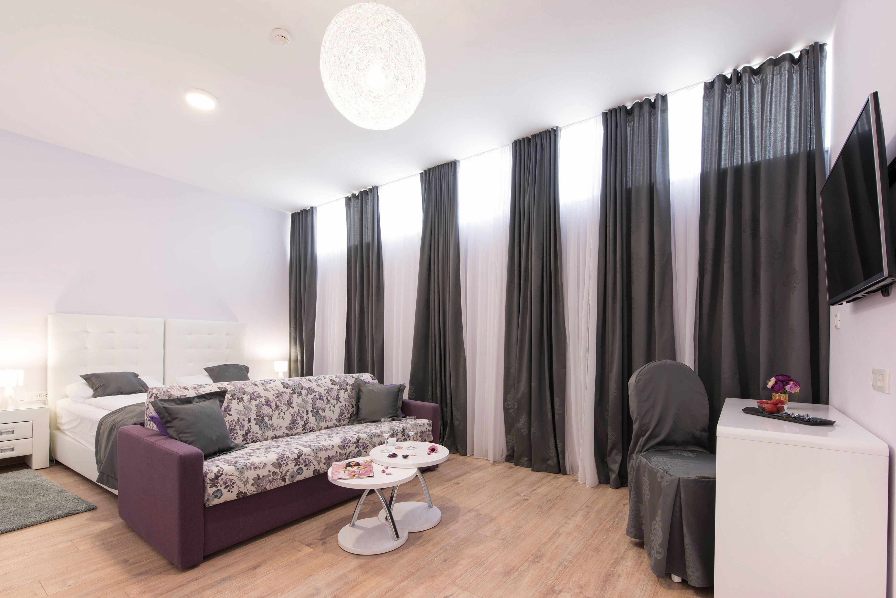 A modern living room with a sofa, a small round table, a flat-screen TV, and a bed with a white headboard.