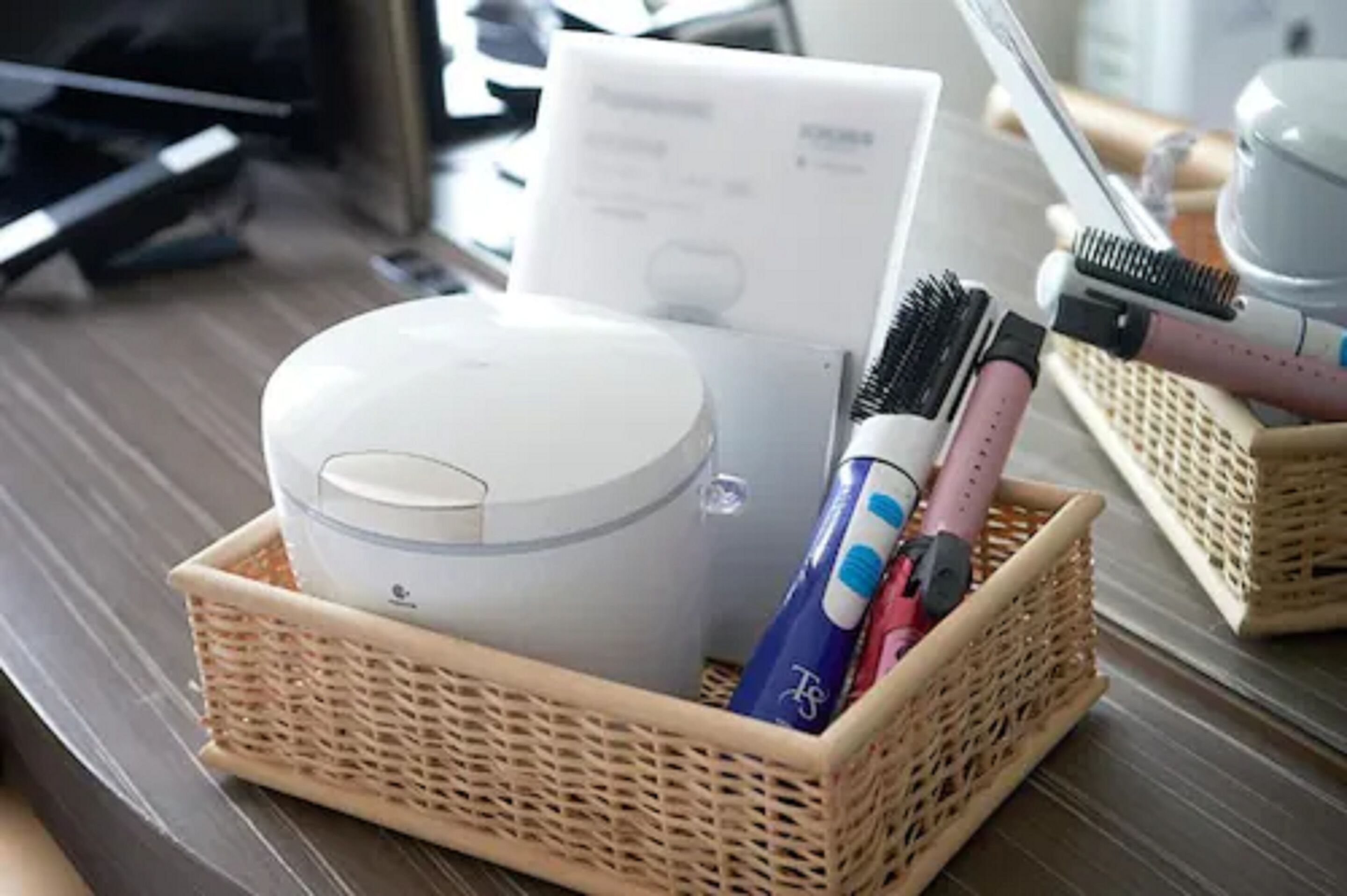 A wicker basket containing a white cylindrical container, a blue and pink hairbrush, and a white box with text.