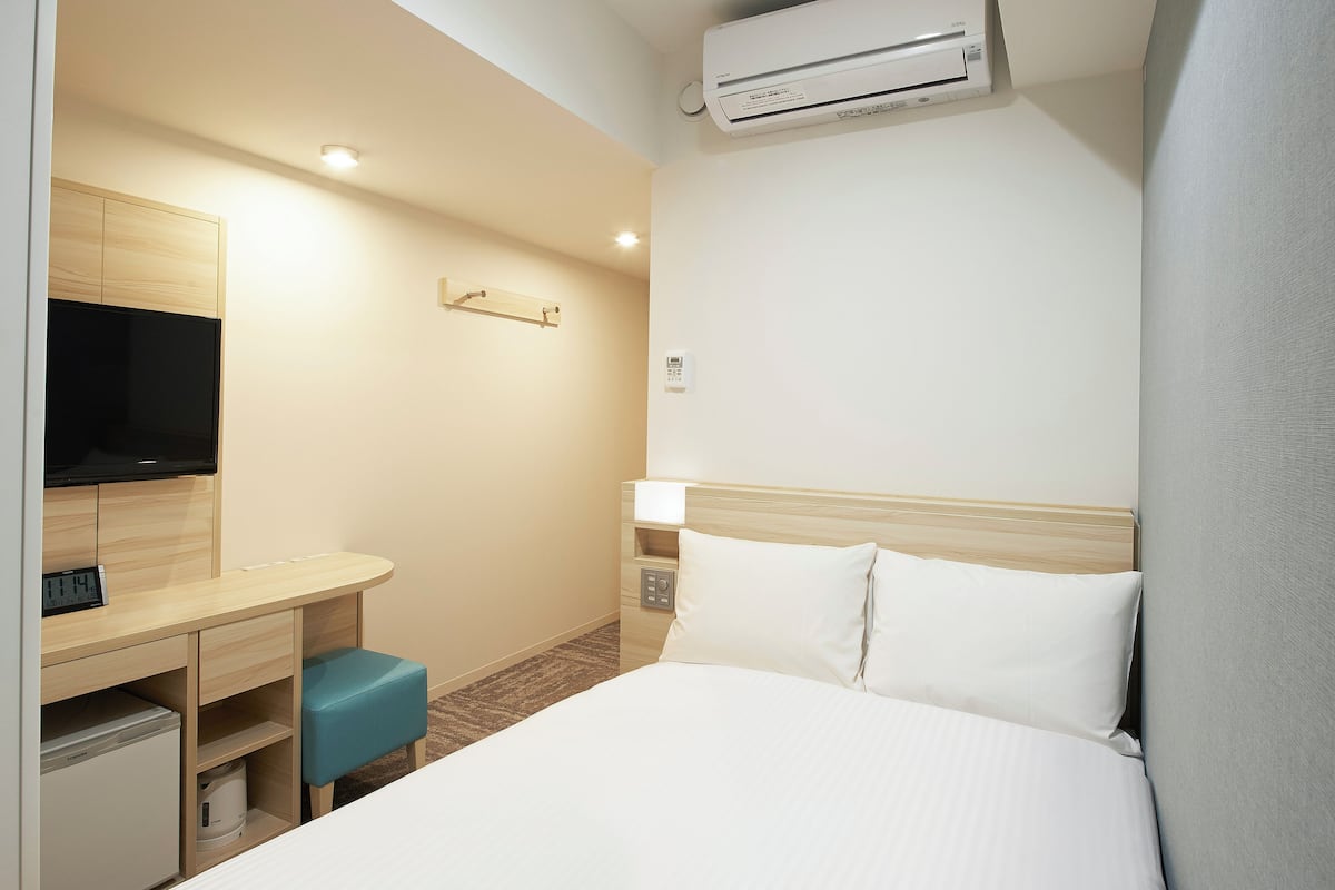 A hotel room with a bed, a desk with a television, a small refrigerator, and an air conditioning unit.