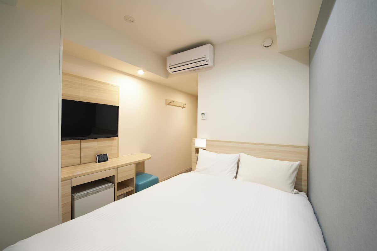 A hotel room with a bed, a desk with a flat-screen TV, a small seating area, and an air conditioning unit.