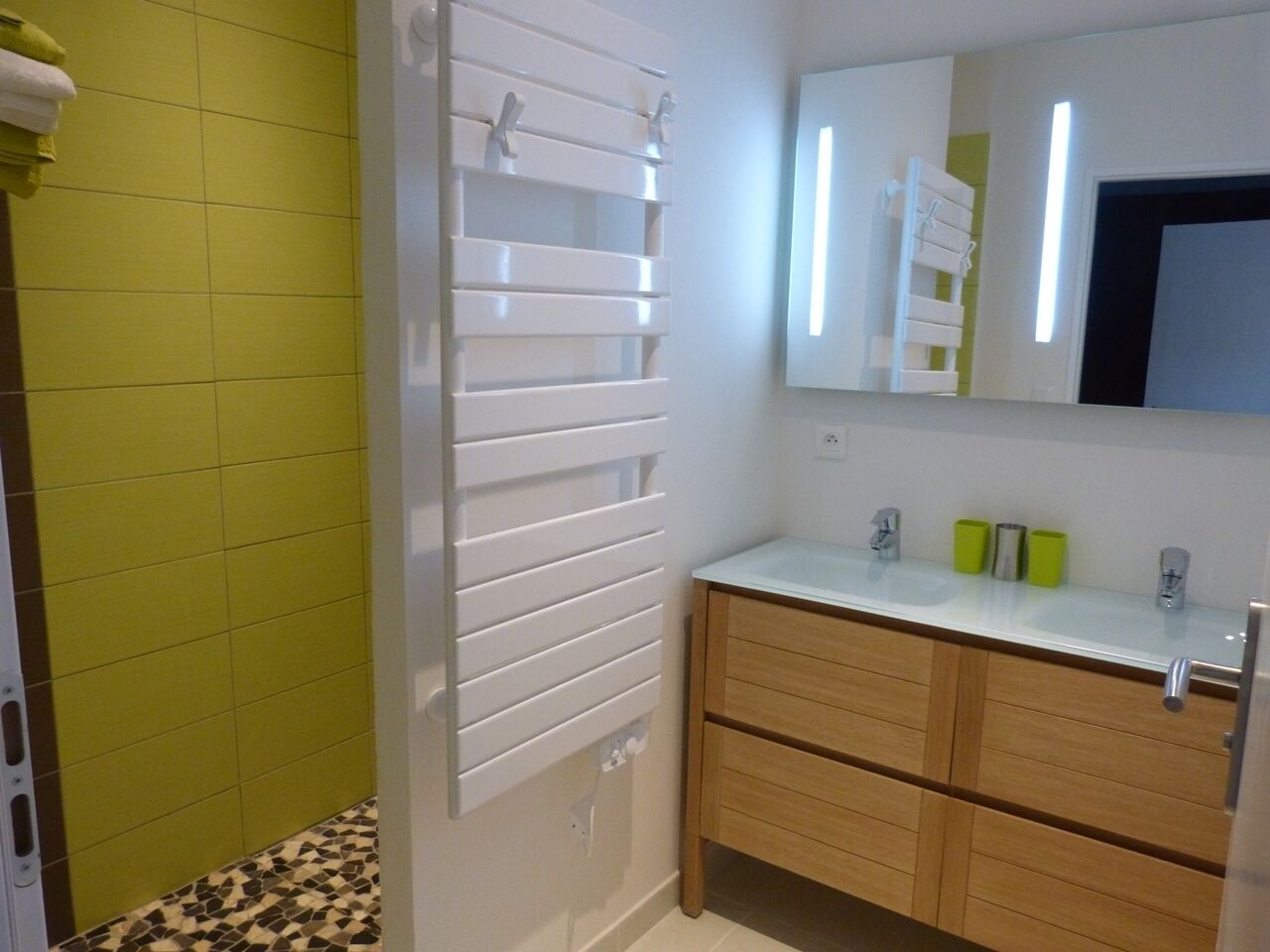 Un baño con pared de azulejos verdes, radiador blanco, tocador de madera con dos cajones y espejo sobre el tocador.