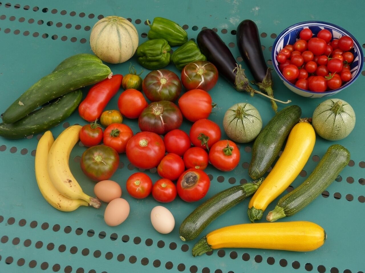 Diversitat de fruites i verdures fresques, com ara tomàquets, pepinos, albergínies i plàtans, disposades sobre una superfície blava amb dibuixos de punts.
